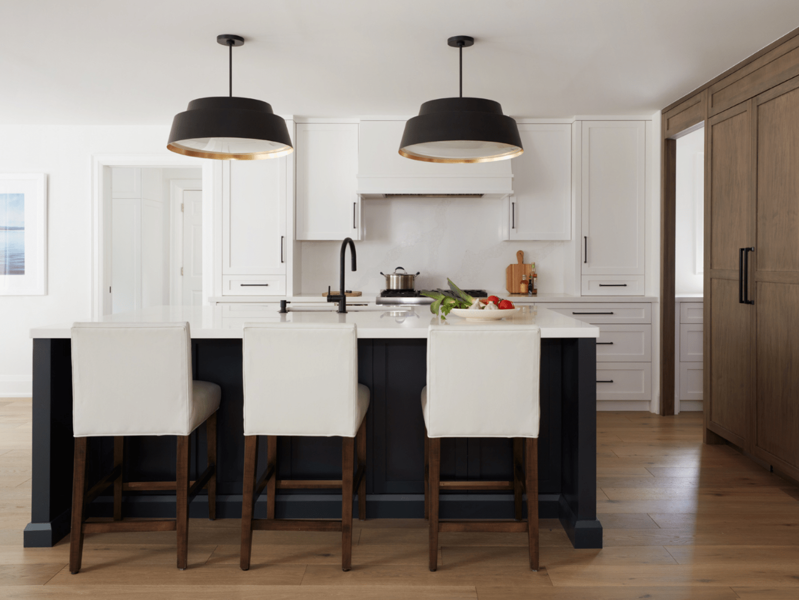 Transitional kitchen with white upper cabinets and range hood, dark painted island, stained maple wood pantry wall